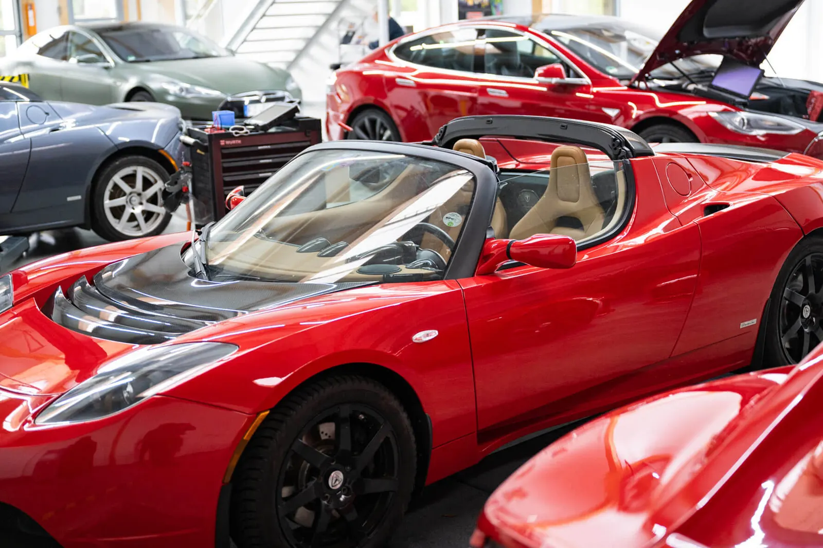A look into the workshop hall: a red Tesla Roadster with its roof off in the foreground, more electric vehicles and a tool trolley behind it.